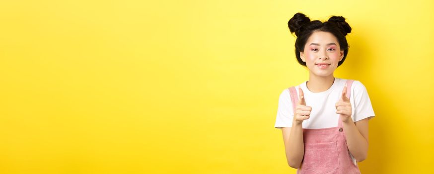 Cheerful teen asian girl pointing fingers at camera and smiling, encourage or invite you, making compliment, praise nice work, standing on yellow background.