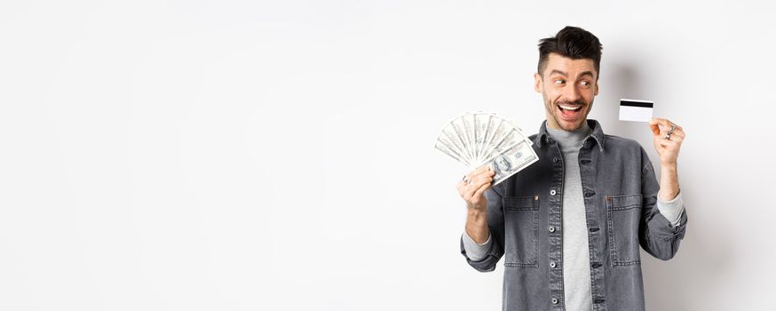 Excited guy holding plastic credit card and dollar bills, standing amused on white background.