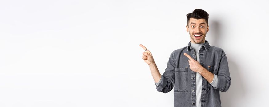 Excited guy with moustache pointing fingers left, showing advertisement and smiling happy, standing on white background.