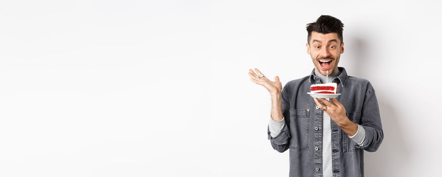 Excited man celebrating birthday, looking happy at bday cake with candle, making wish, standing happy against white background.