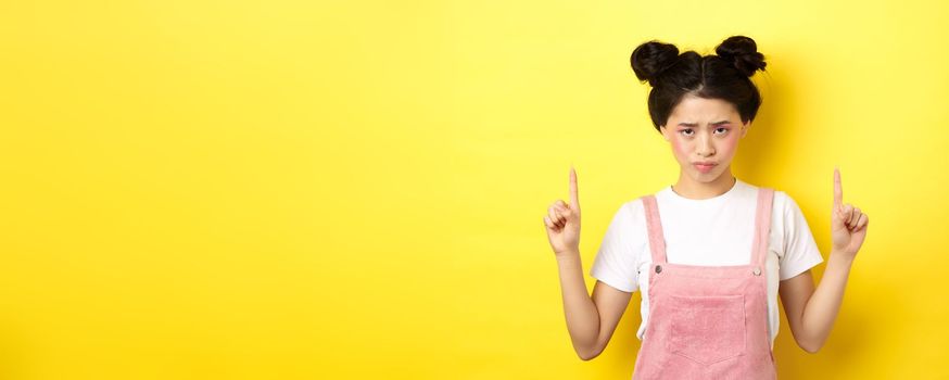 Sad and gloomy asian woman frowning, pointing fingers up and complaining on unfair thing, standing upset on yellow background.