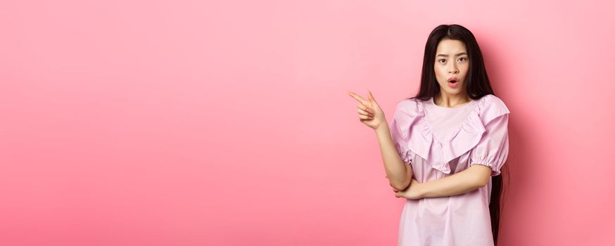 Shocked asian teen girl in dress pointing finger left, gasping wondered and look curious at camera, asking question about promo, standing on pink background.