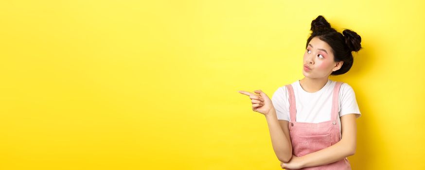 Pensive asian woman with beauty makeup, pointing and looking left with curious face, interested in advertisement, standing on yellow background.