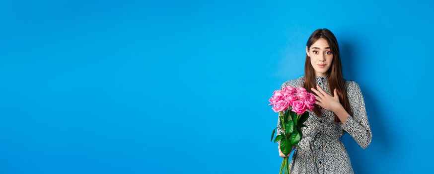 Valentines day concept. Touched and thankful pretty girl, holding hand on heart and smiling grateful for gift, holding bouquet of roses, standing on blue background.