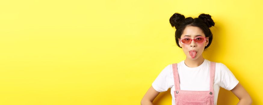 Summer and fashion concept. Silly asian teen girl showing tongue, wearing sunglasses, standing on yellow background.