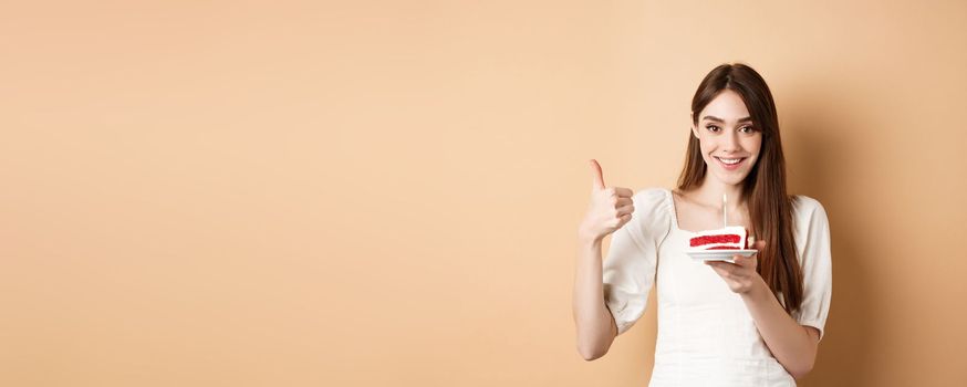 Beautiful birthday girl making wish on cake, showing thumb up and smiling happy, enjoying b-day party, standing on beige background.