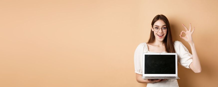 E-commerce. Excited young woman in glasses showing okay sign and laptop screen, recommending internet promo, standing on beige background.