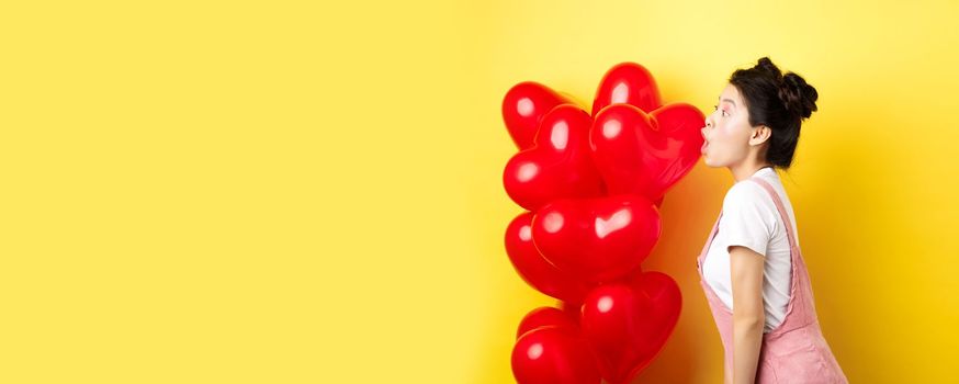 Valentines day and relationship concept. Profile of young asian woman scream of surprise, say wow and looking left amazed, standing near red balloons, yellow background.