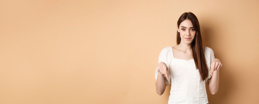 Smiling tender girl in dress pointing fingers down, showing advertisement on empty space, beige background.