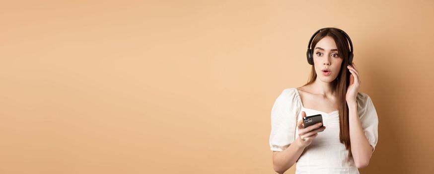 Wow awesome tunes. Excited young girl amazed with new song, listening music in headphones and look impressed, holding cell phone, beige background.