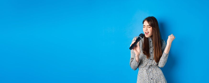 Pretty natural girl in dress singing songs in microphone, holding mic and looking passionate while performing, standing on blue background.