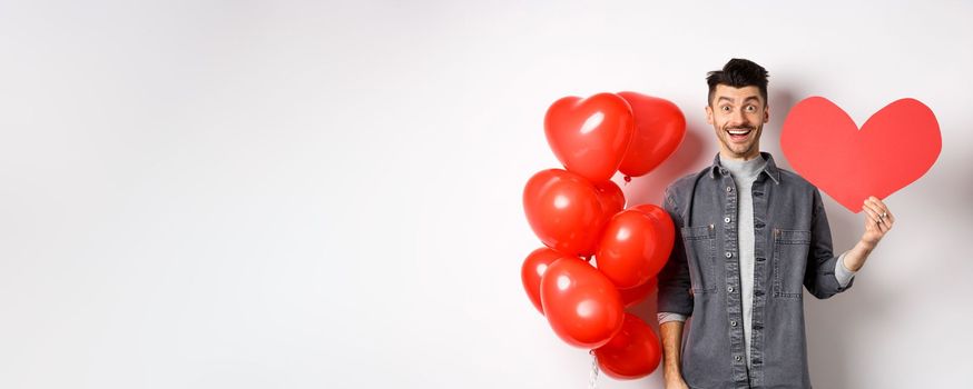 Valentines day and love concept. Cheerful funny guy showing heart cutout, standing near romantic balloons and smiling excited at camera, white background.