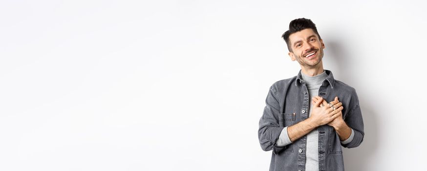 Romantic guy hold hands on heart and smiling, thanking you, looking pleased and grateful, standing on white background.