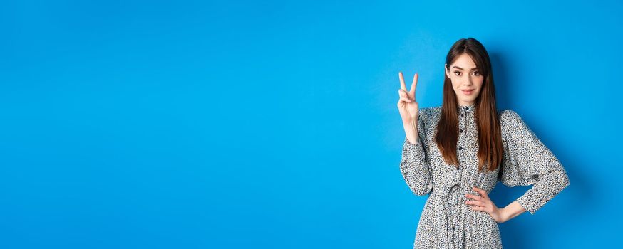 Young beautiful girl in dress with natural long hair, showing number two with fingers and smiling, standing against blue background.