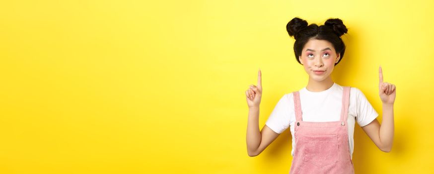 Glamour asian girl with beauty makeup, pointing fingers up, showing top advertisement, standing on yellow background.