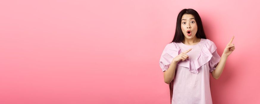 Excited asian woman look amazed, pointing right and gasping fascinated, checking out advertisement, standing on pink background.