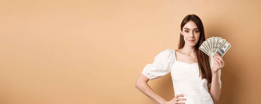 Confident smiling woman showing dollar bills, earn money and look satisfied, standing on beige background with cash.