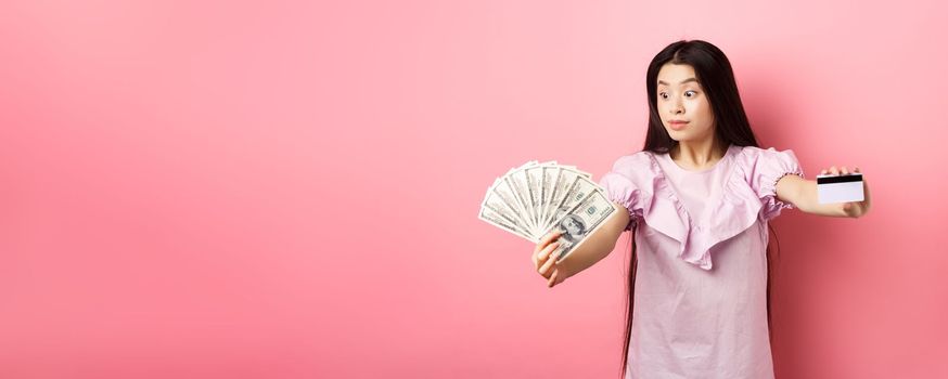 Excited teen asian girl stretch out hands with dollar bills and plastic credit card, look at money with amazement, standing on pink background.