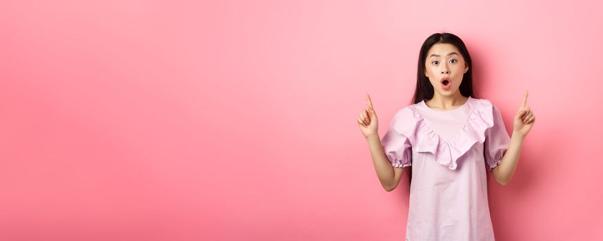 Excited asian teen girl pointing fingers up, saying wow and showing promo deal, standing in dress against pink background.