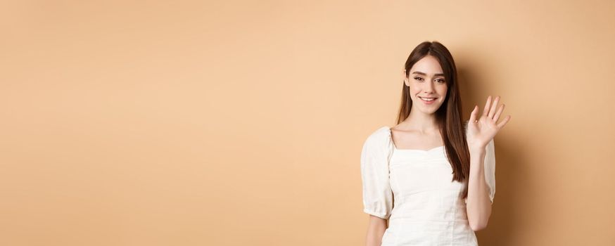 Friendly smiling woman say hello, waiving hand to greet you, standing cheerful on beige background.