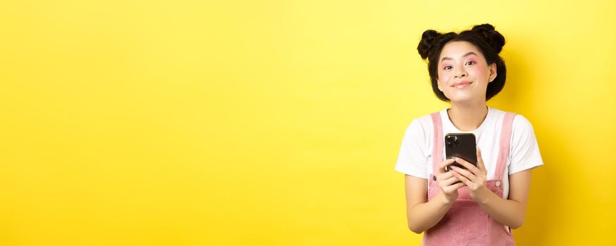 Cute stylish asian girl using mobile phone, wearing glamour pink makeup and summer clothes, standing on yellow background.