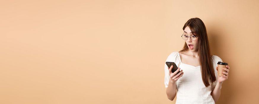 Surprised girl reading exciting news on cell phone, drop jaw amazed, standing with coffee cup on beige background.