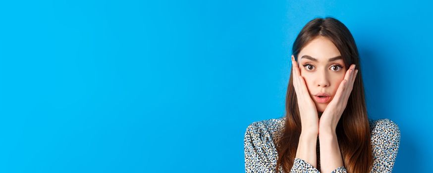 Close-up portrait of surprised cute woman holding hands on cheeks and gasping, stare at camera shocked, standing against blue background.