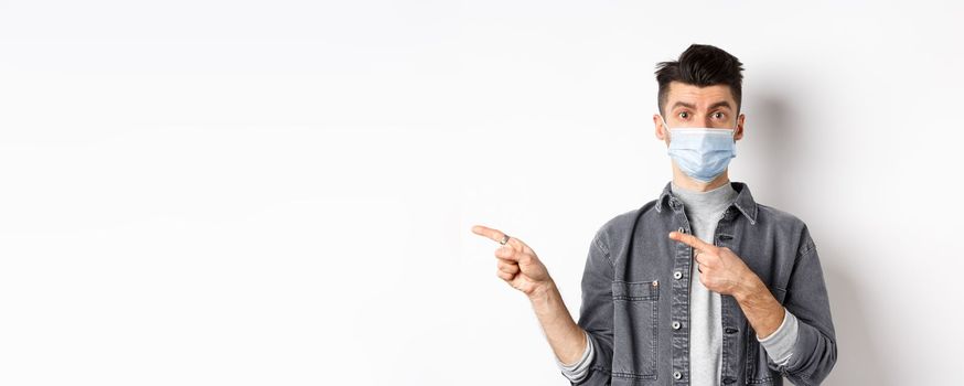 Pandemic lifestyle, healthcare and medicine concept. Young guy in face mask inviting look here, pointing fingers left at logo, standing on white background.