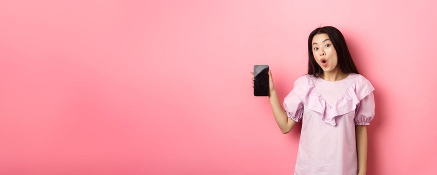 Online shopping. Excited asian woman say wow, showing empty smartphone screen and looking amazed at camera, standing against pink background.