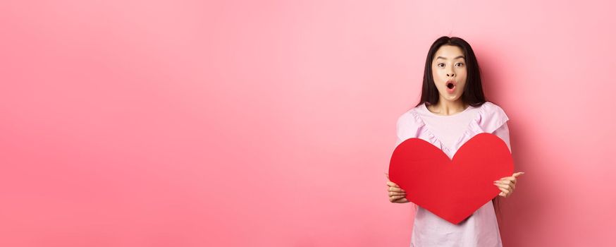 Valentines day concept. Surprised asian teen girl say wow, showing big red heart card, receive gift from secret admirer or lover, standing in romantic dress on pink background.
