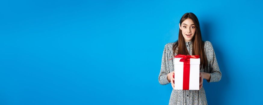 Valentines day concept. Surprised happy girl receiving romantic gift and looking thankful at camera, standing on blue background.