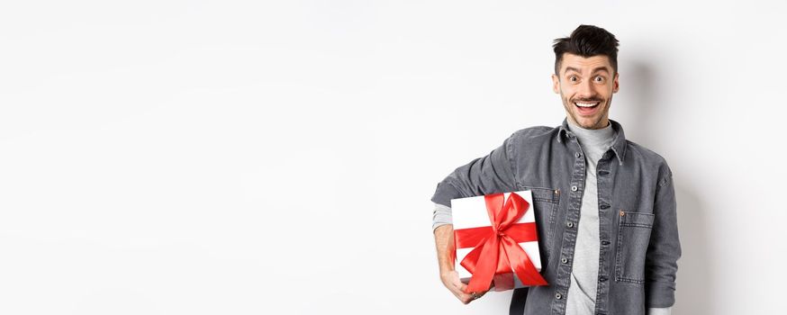 Happy Valentines day. Surprised and happy guy holding gift box and look at camera, smiling amazed, celebrating holiday, bring present on romantic date, white background.