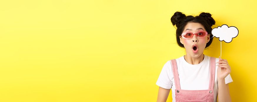 Summer and fashion concept. Excited party girl in sunglasses, holding comment cloud party mask and gasping amazed, standing impressed on yellow background.