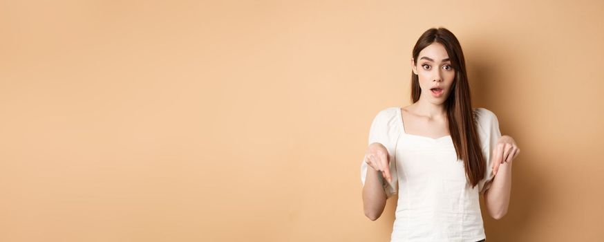 Wow look there. Amazed young woman in white dress pointing fingers down, standing in awe with dropped jaw and popped eyes against beige background.