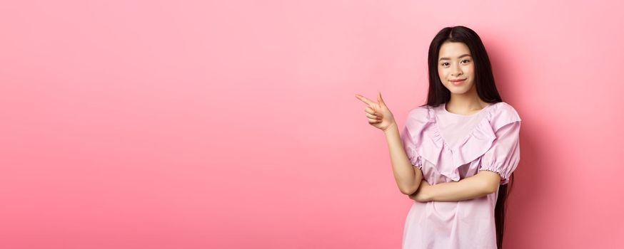 Beautiful teenage asian girl pointing finger left, smiling confident, showing advertisement on pink background.