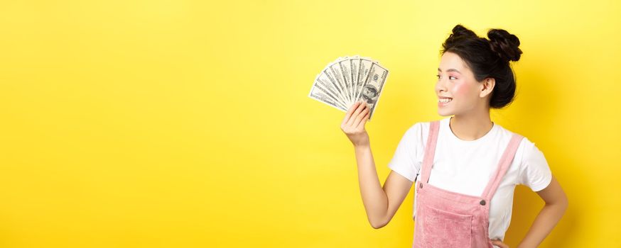 Shopping. Rich and stylish asian female model showing money, looking at dollar bills with pleased smile, standing on yellow background.