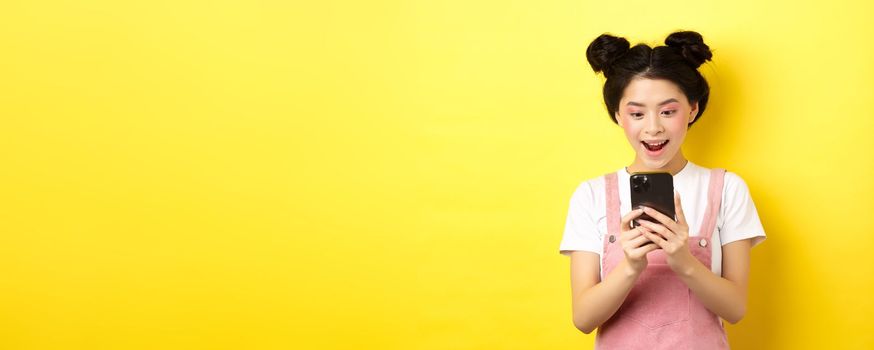 Pretty asian girl looking excited at screen, reading message on phone and smiling happy, standing in summer clothes on yellow background.