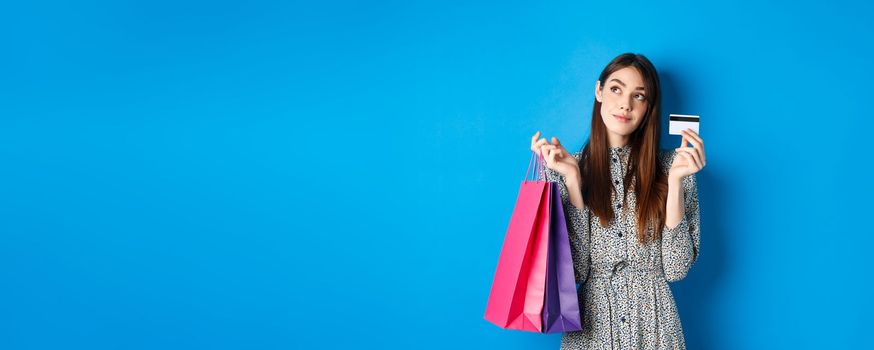 Dreamy girl looking at logo and showing plastic credit card, holding shopping bags, standing on blue background.