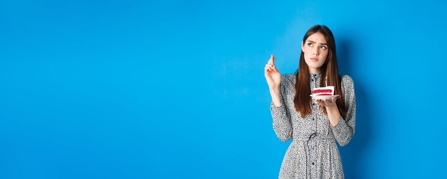 Thoughtful birthday girl making wish, cross fingers good luck, holding cake and looking at upper left corner, blue background.