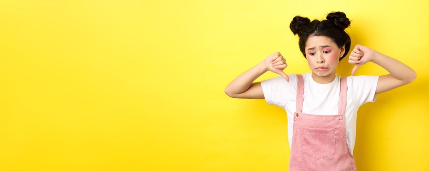 Asian beauty girl cringe from something disgusting, look away and showing thumbs-down to express dislike, negative emotions, standing in summer clothes on yellow background.