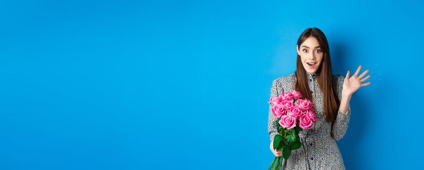 Valentines day concept. Surprised pretty girl holding bouquet of flowers from lover, looking amazed and happy at camera, standing on blue background.