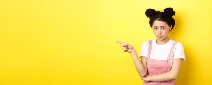 Sad and gloomy asian girl with glamour makeup, frowning unfair and pointing left at logo, looking disappointed, standing on yellow background.