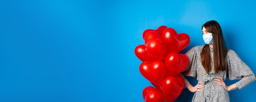 Covid-19 and Valentines day. Young bothered girl in medical mask and dress, looking at heart balloons, waiting on date, standing blue background.