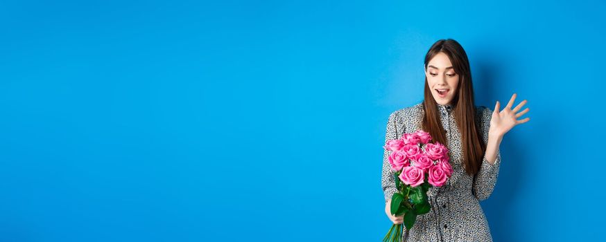 Valentines day concept. Image of attractive young woman gasping amazed, receive surprise flowers, standing on blue background.
