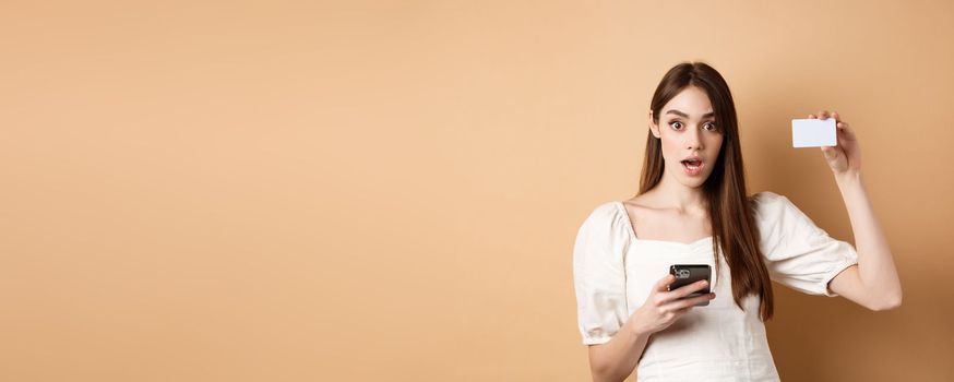 Excited woman showing plastic credit card and using mobile phone app, drop jaw and gasping amazed, checking out bank offer, beige background.