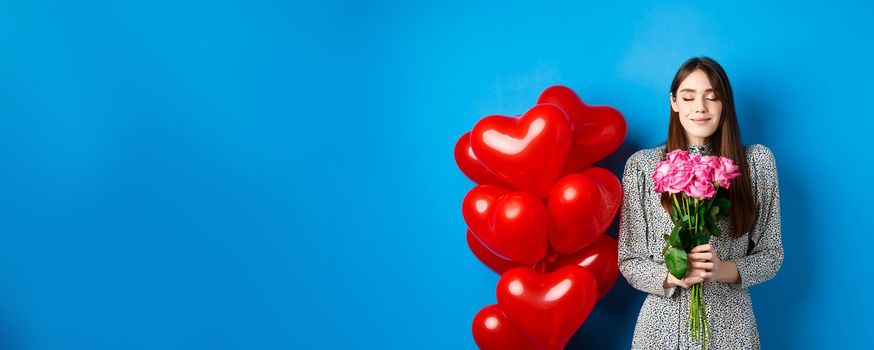 Valentines day. Romantic pretty woman close eyes and smelling beautiful flowers, standing near heart balloons, blue background.