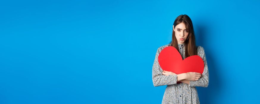 Valentines day. Sad and gloomy caucasian girl pucker lips and looking disappointed, holding big red heart cutout, standing on blue background.