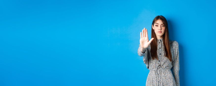 Serious and confident woman with long hair and dress, stretch out hand and say no, make stop gesture, prohibit bad action, standing on blue background.