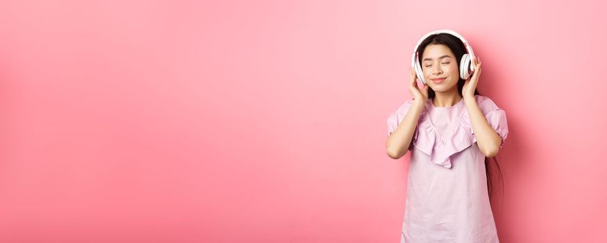 Beautiful asian girl close eyes while listening music in headphones, enjoying soft sound, standing against pink background.