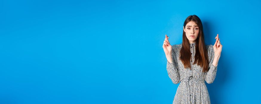 Nervous young woman in dress cross fingers and biting lip worried, praying or making wish, standing on blue background.